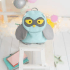 Crochetts - MOCHILA BUHO Una linda mochila MOCHILA BUHO de Crochetts se encuentra sobre un pequeño taburete blanco, con un diseño de color azul claro con alas grises y un pico amarillo. Los globos brillantes y las luces de cadena en el fondo le dan un toque festivo, lo que la hace perfecta para preparar una celebración junto con chupetes de bebé asequibles.