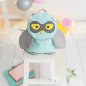 Crochetts - MOCHILA BUHO Una linda mochila MOCHILA BUHO de Crochetts se encuentra sobre un pequeño taburete blanco, con un diseño de color azul claro con alas grises y un pico amarillo. Los globos brillantes y las luces de cadena en el fondo le dan un toque festivo, lo que la hace perfecta para preparar una celebración junto con chupetes de bebé asequibles.