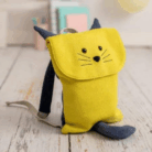 Crochetts - MOCHILA LOBITO Una MOCHILA LOBITO de Crochetts, que se asemeja a un gato con cuerpo amarillo y orejas y cola azules, reposa sobre un piso de madera clara. Los globos de colores en el fondo sugieren celebraciones. ¡Recuerde aprovechar ofertas en parafarmacia infantil mientras se prepara para días llenos de diversión!