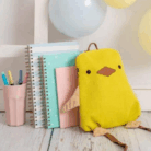 Crochetts - MOCHILA POLLITO Una encantadora MOCHILA POLLITO de Crochetts con forma de pájaro descansa junto a cuadernos de espiral y una taza rosa con bolígrafos. Los globos pastel realzan la festiva escena del suelo de madera clara, perfecta para los recuerdos mientras aprovechas las ofertas en parafarmacia infantil para las necesidades de tu pequeño.