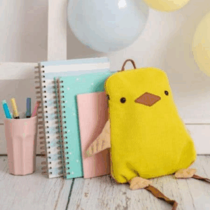 Crochetts - MOCHILA POLLITO Una encantadora MOCHILA POLLITO de Crochetts con forma de pájaro descansa junto a cuadernos de espiral y una taza rosa con bolígrafos. Los globos pastel realzan la festiva escena del suelo de madera clara, perfecta para los recuerdos mientras aprovechas las ofertas en parafarmacia infantil para las necesidades de tu pequeño.